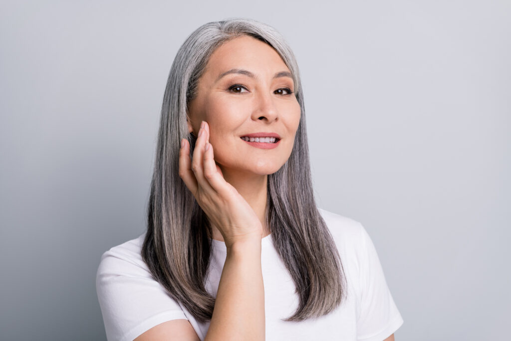 Photo portrait of senior woman with grey hair touching cheek wearing white t-shirt smiling isolated on grey color background.