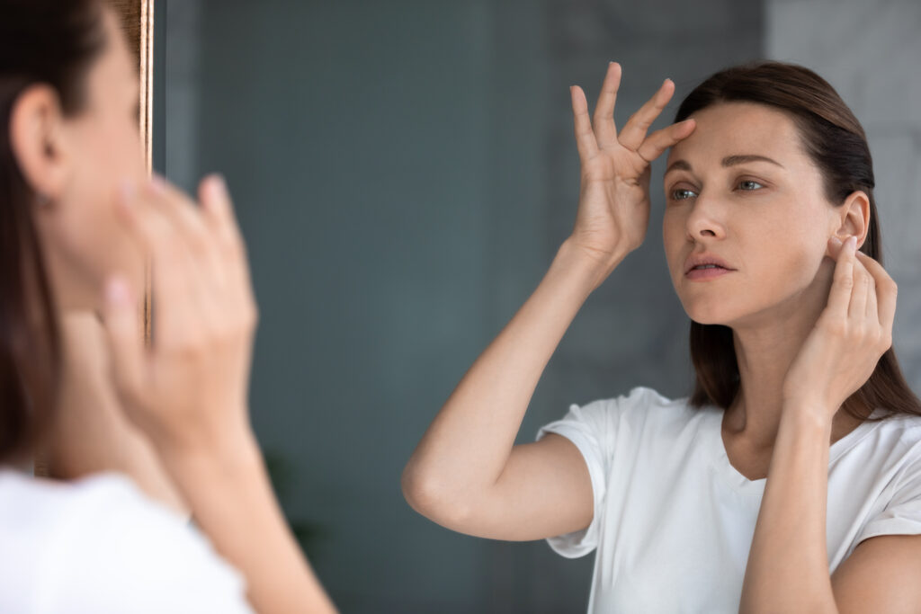 Anxious woman looking in mirror, standing in bathroom, upset attractive female touching forehead with finger, confused about wrinkles or acne, thinking about face lifting procedure or spa