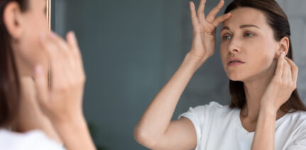 Anxious woman looking in mirror, standing in bathroom, upset attractive female touching forehead with finger, confused about wrinkles or acne, thinking about face lifting procedure or spa