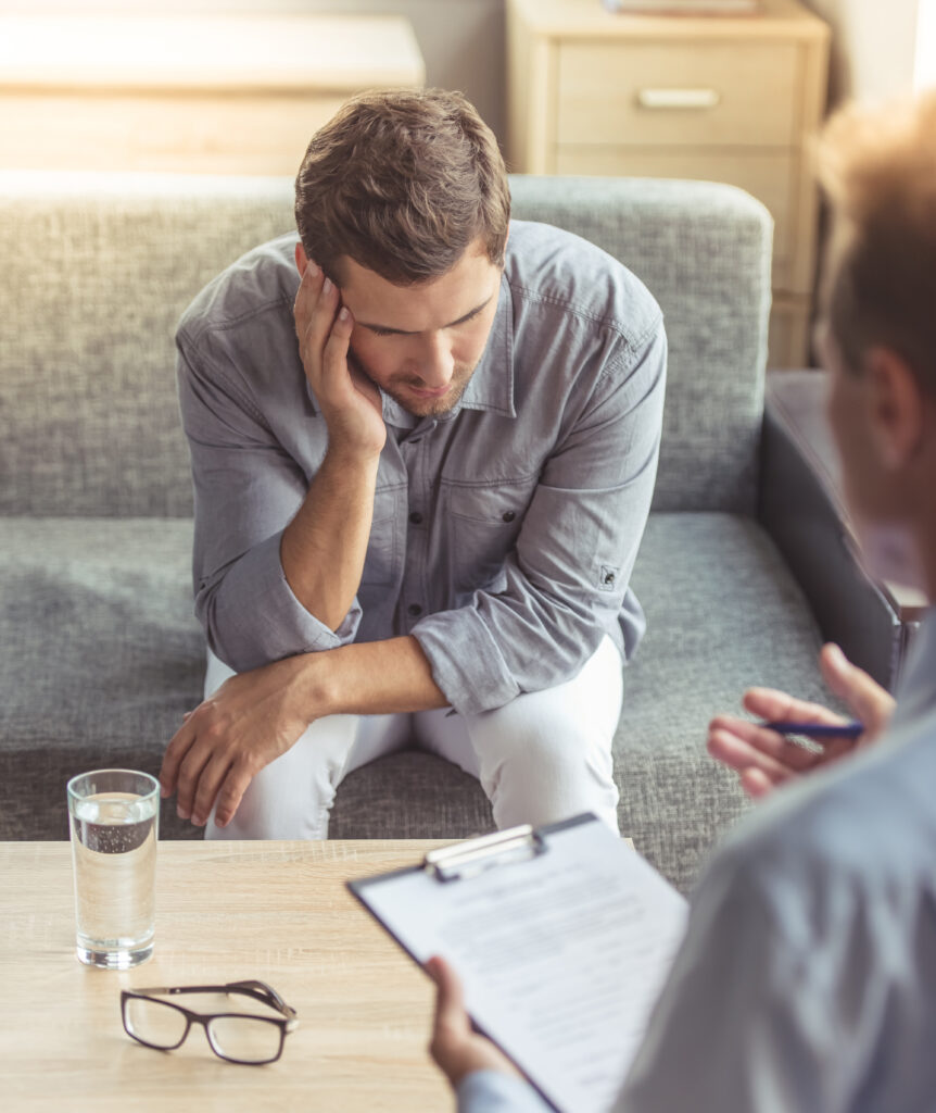 Handsome depressed man in casual clothes is leaning on his knees and telling about his problems while sitting on the couch at the psychotherapist